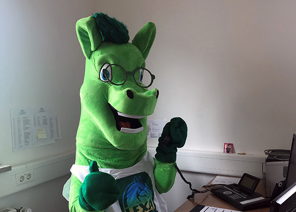 Grünes Maskottchen am Telefon Ein grünes Maskottchen mit Brille und einem Lächeln, das am Telefon steht und Daumen hoch zeigt.
