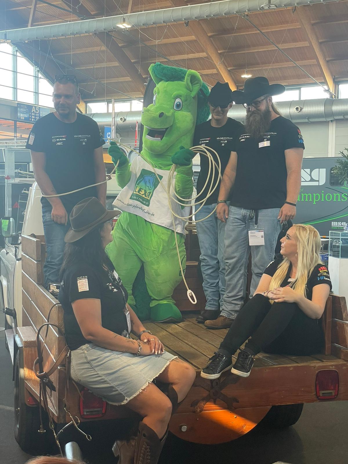 Grünes Maskottchen auf einem Pickup mit Personen Eine grüne Maskottchenfigur steht auf einem Pickup, umgeben von mehreren Personen in Cowboyhüten und T-Shirts.