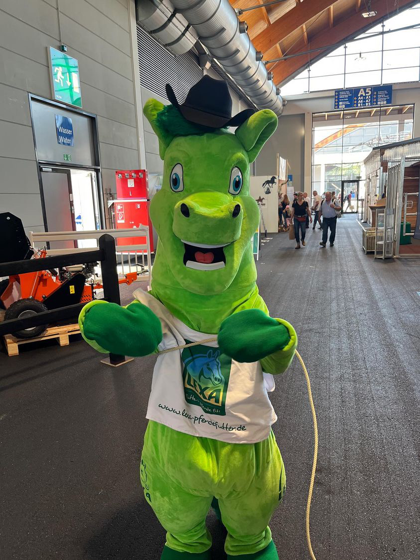 Grünes Maskottchen mit Cowboyhut Ein grünes Maskottchen in einem Cowboyhut, das mit den Händen eine Lasso-Geste macht.
